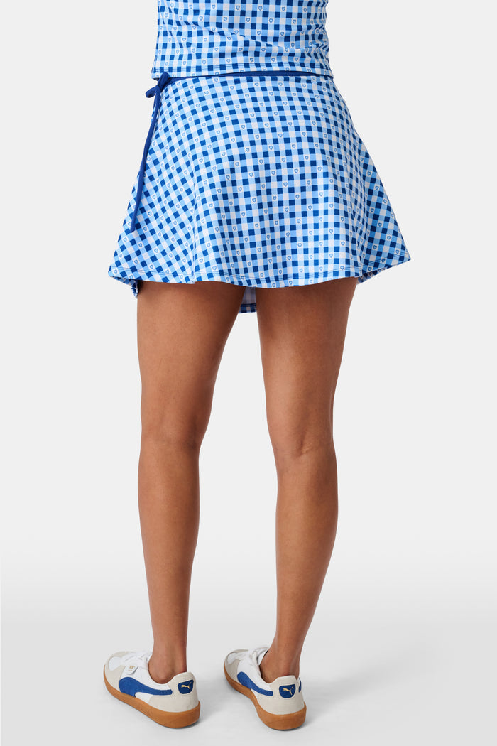 Wearing the Roller Rabbit Gingham Hearts Coco Wrap Skirt, a woman stands next to a net outdoors, surrounded by green grass and trees.