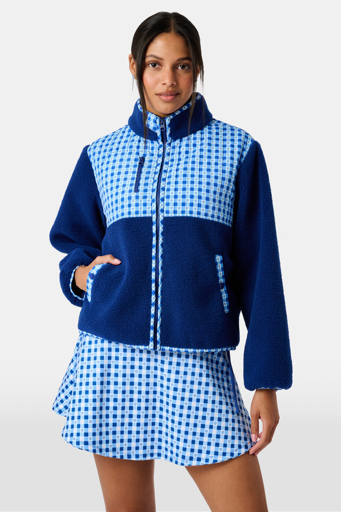 A woman wearing the Roller Rabbit Gingham Hearts Marina Fleece Jacket stands by a sports net outdoors, holding a patterned tote and a water bottle.