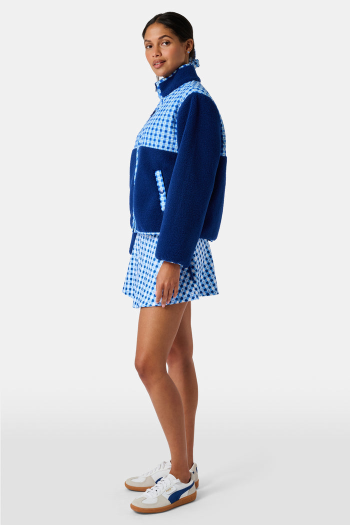 A woman wearing the Roller Rabbit Gingham Hearts Marina Fleece Jacket stands by a sports net outdoors, holding a patterned tote and a water bottle.