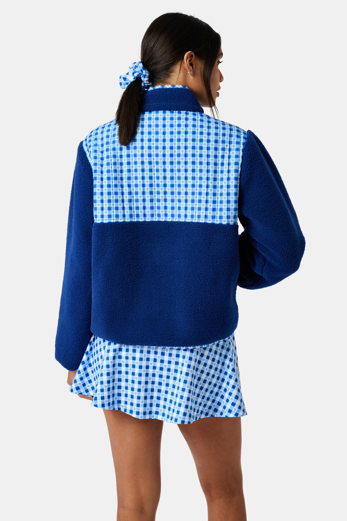 A woman wearing the Roller Rabbit Gingham Hearts Marina Fleece Jacket stands by a sports net outdoors, holding a patterned tote and a water bottle.