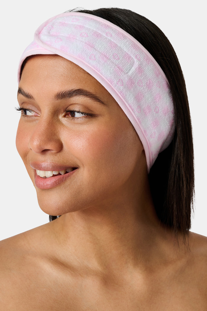 The Roller Rabbit Gingham Hearts Spa Headband is crafted from 100 cotton terrycloth and showcases a pink and white checkered pattern accented with small hearts. It features a convenient hook-and-loop fastener and is displayed laid flat on a white background.