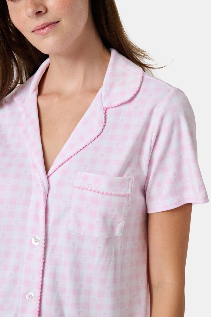 The Gingham Hearts Wyatt Polo Pajamas by Roller Rabbit are a pink and white gingham short-sleeve pajama set, featuring a button-up shirt with chest pocket and matching shorts with a ruffled hem, all crafted from soft Pima cotton. This set is shown on a white background.