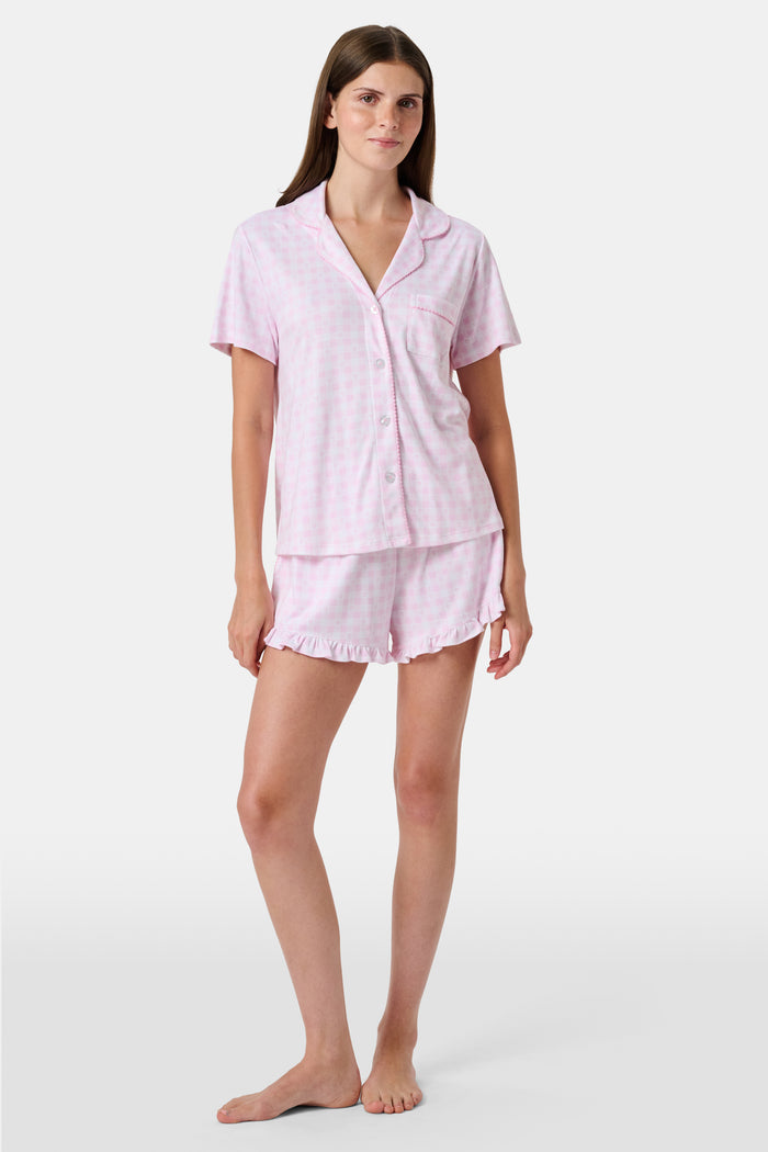 The Gingham Hearts Wyatt Polo Pajamas by Roller Rabbit are a pink and white gingham short-sleeve pajama set, featuring a button-up shirt with chest pocket and matching shorts with a ruffled hem, all crafted from soft Pima cotton. This set is shown on a white background.