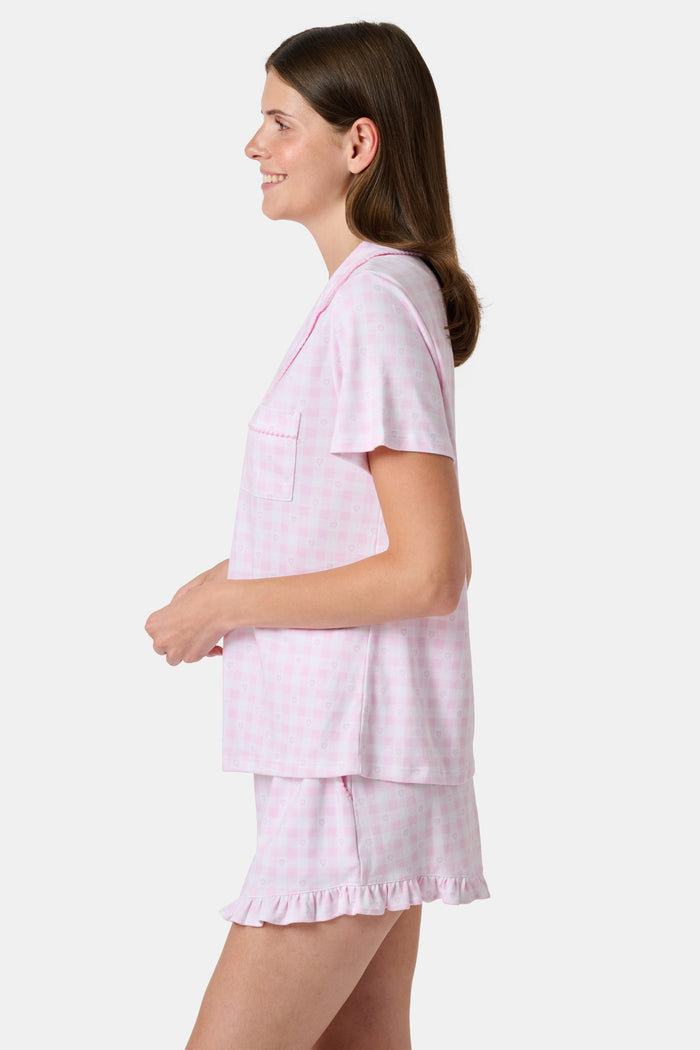 The Gingham Hearts Wyatt Polo Pajamas by Roller Rabbit are a pink and white gingham short-sleeve pajama set, featuring a button-up shirt with chest pocket and matching shorts with a ruffled hem, all crafted from soft Pima cotton. This set is shown on a white background.