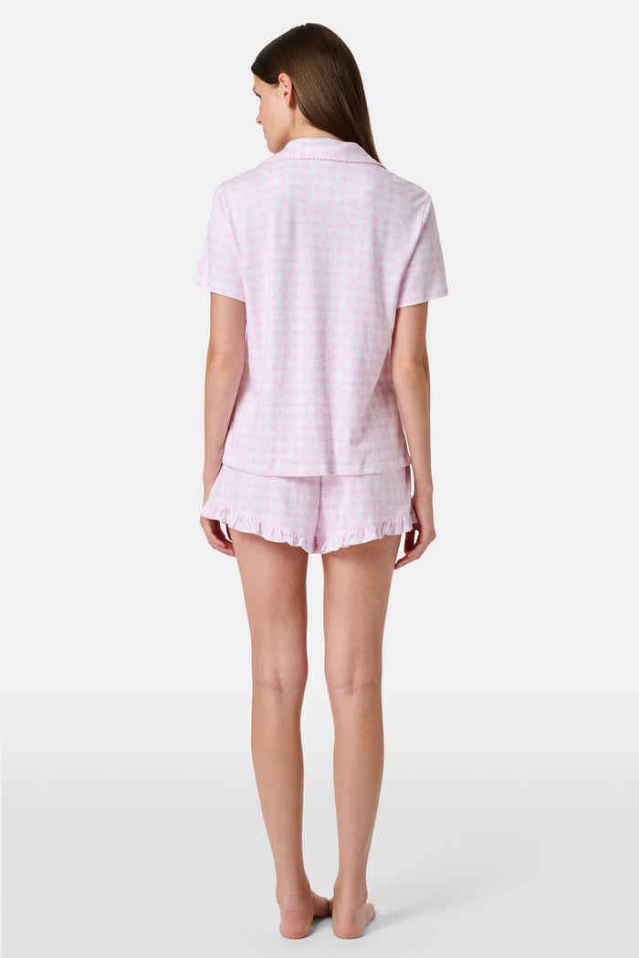 The Gingham Hearts Wyatt Polo Pajamas by Roller Rabbit are a pink and white gingham short-sleeve pajama set, featuring a button-up shirt with chest pocket and matching shorts with a ruffled hem, all crafted from soft Pima cotton. This set is shown on a white background.