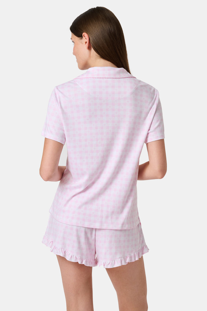 The Gingham Hearts Wyatt Polo Pajamas by Roller Rabbit are a pink and white gingham short-sleeve pajama set, featuring a button-up shirt with chest pocket and matching shorts with a ruffled hem, all crafted from soft Pima cotton. This set is shown on a white background.