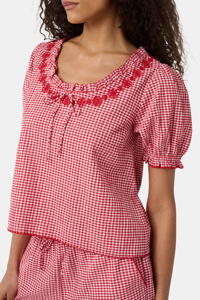 A woman wears the Roller Rabbit Gingham Quincy Top, a cotton blouse with puffed sleeves and red embroidery at the neckline. Only her upper body is visible.