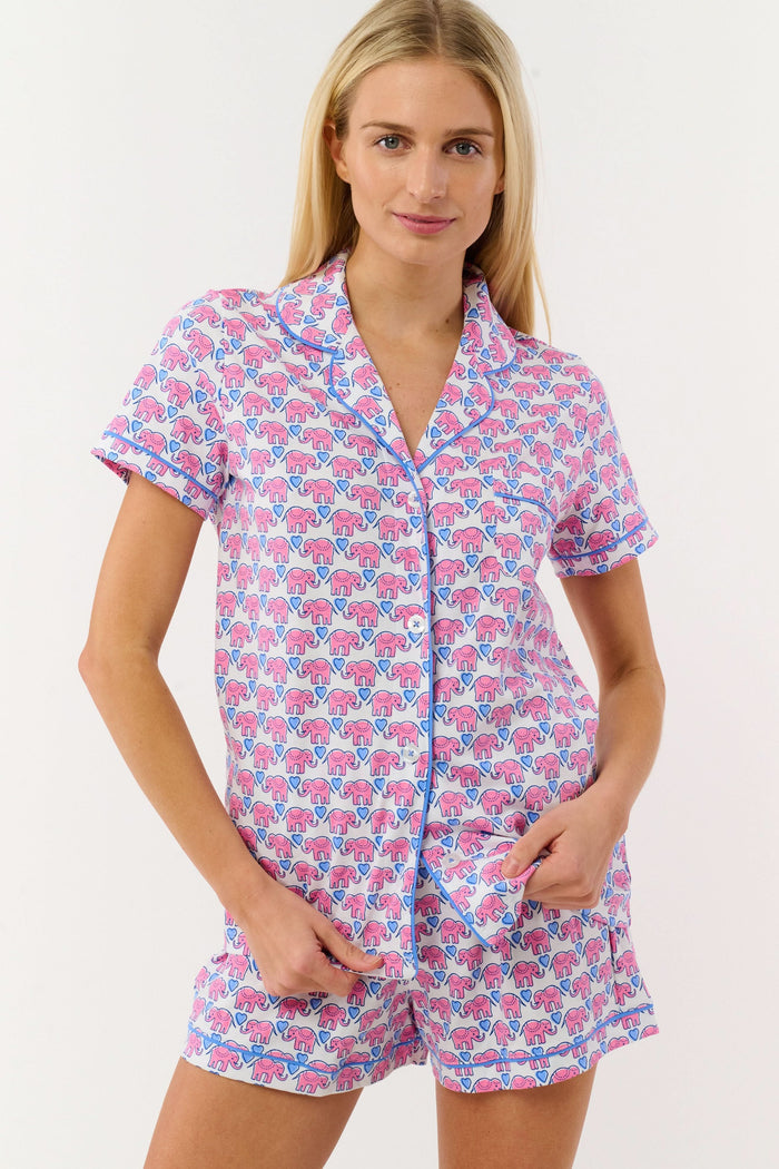 A woman with long blonde hair smiles slightly while wearing Roller Rabbit's Hathi Hearts Polo Pajamas, a soft Pima cotton matching set with pink and blue elephant print, short-sleeve button-up top, shorts, and blue piping.