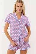 A woman with long blonde hair smiles slightly while wearing Roller Rabbit's Hathi Hearts Polo Pajamas, a soft Pima cotton matching set with pink and blue elephant print, short-sleeve button-up top, shorts, and blue piping.