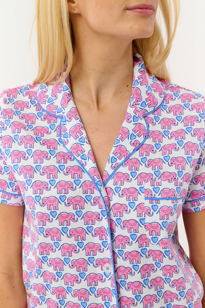 A woman with long blonde hair smiles slightly while wearing Roller Rabbit's Hathi Hearts Polo Pajamas, a soft Pima cotton matching set with pink and blue elephant print, short-sleeve button-up top, shorts, and blue piping.