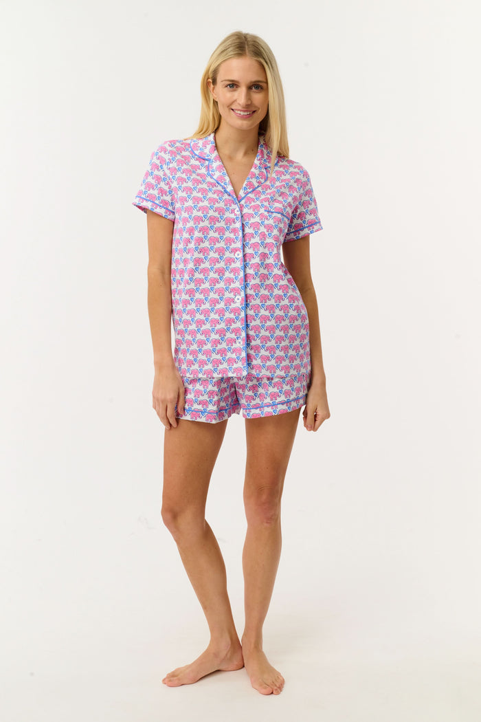 A woman with long blonde hair smiles slightly while wearing Roller Rabbit's Hathi Hearts Polo Pajamas, a soft Pima cotton matching set with pink and blue elephant print, short-sleeve button-up top, shorts, and blue piping.