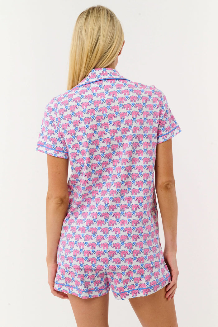 A woman with long blonde hair smiles slightly while wearing Roller Rabbit's Hathi Hearts Polo Pajamas, a soft Pima cotton matching set with pink and blue elephant print, short-sleeve button-up top, shorts, and blue piping.