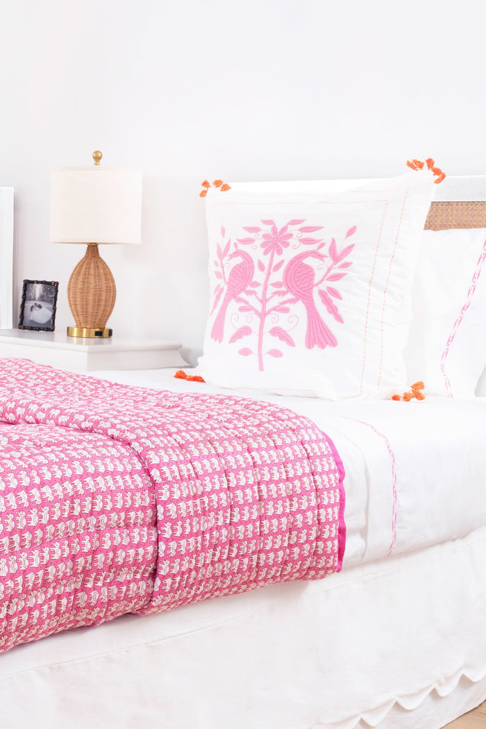 A neatly made bed showcases the Roller Rabbit Hathi Quilt, paired with a white comforter, pink patterned blanket, and a pillow adorned with pink birds and flowers. In the background, a wooden lamp and framed photo rest on a white nightstand.