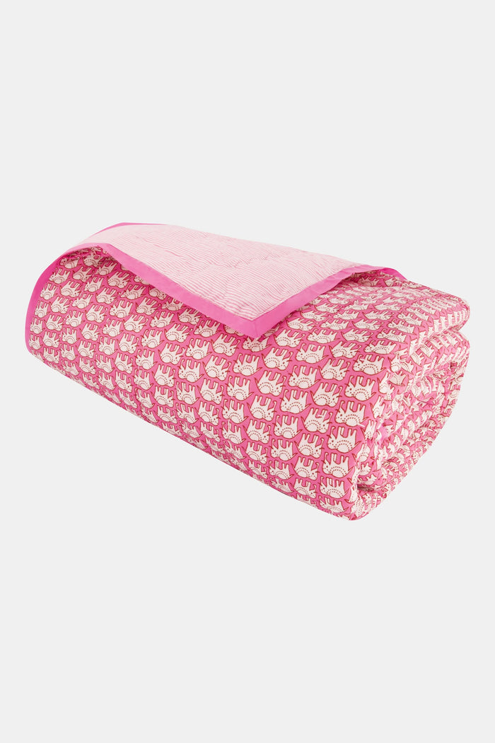 A neatly made bed showcases the Roller Rabbit Hathi Quilt, paired with a white comforter, pink patterned blanket, and a pillow adorned with pink birds and flowers. In the background, a wooden lamp and framed photo rest on a white nightstand.
