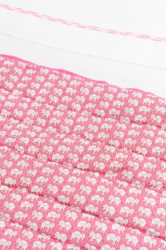A neatly made bed showcases the Roller Rabbit Hathi Quilt, paired with a white comforter, pink patterned blanket, and a pillow adorned with pink birds and flowers. In the background, a wooden lamp and framed photo rest on a white nightstand.
