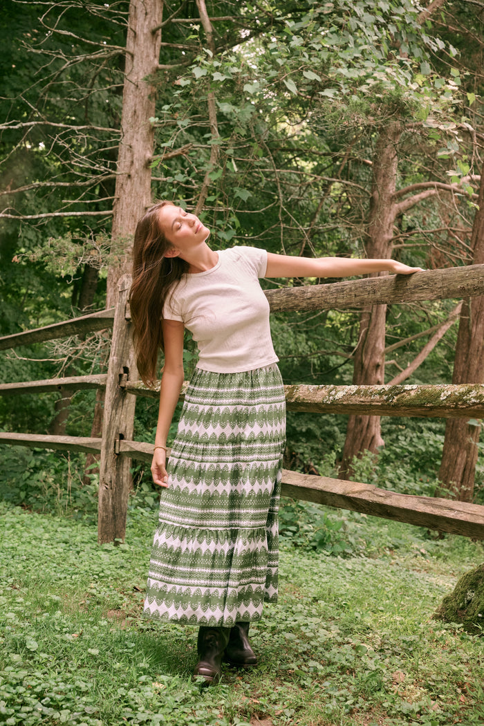 A person wearing the Roller Rabbit Hayam Carlisa Skirt in an olive and white print, paired with a cream knit top and brown boots, stands against a plain white background.