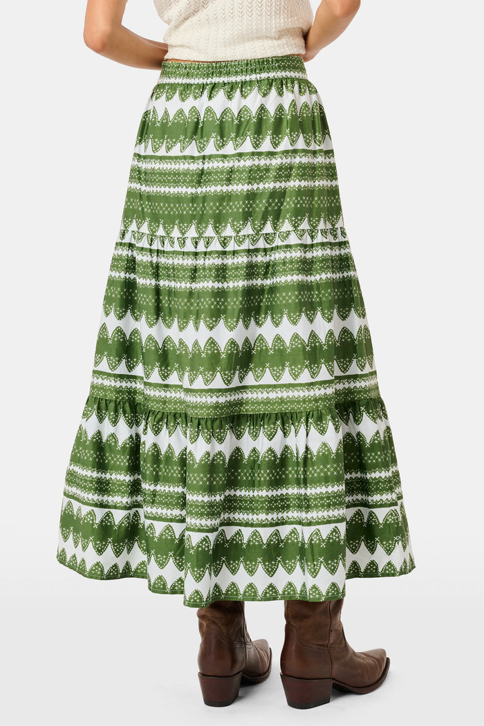A person wearing the Roller Rabbit Hayam Carlisa Skirt in an olive and white print, paired with a cream knit top and brown boots, stands against a plain white background.