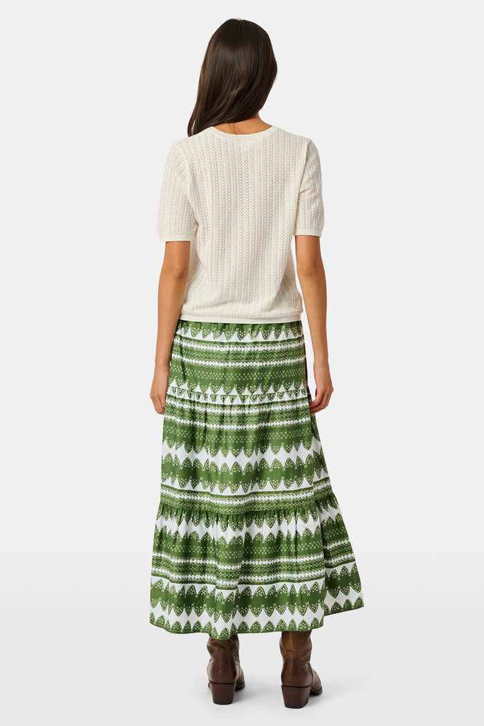 A person wearing the Roller Rabbit Hayam Carlisa Skirt in an olive and white print, paired with a cream knit top and brown boots, stands against a plain white background.