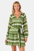 A woman outdoors wears the Hayam Monroe Dress by Roller Rabbit—an olive and white long-sleeved dress with a matching belt. She walks with another person, with trees and a wooden fence in the background.