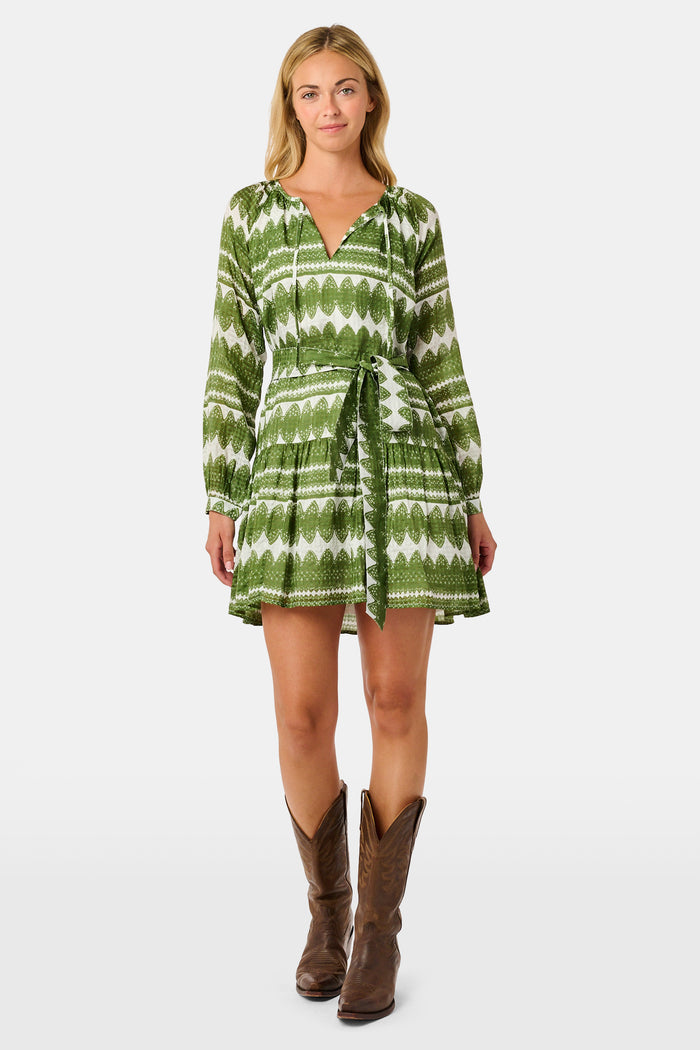 A woman outdoors wears the Hayam Monroe Dress by Roller Rabbit—an olive and white long-sleeved dress with a matching belt. She walks with another person, with trees and a wooden fence in the background.