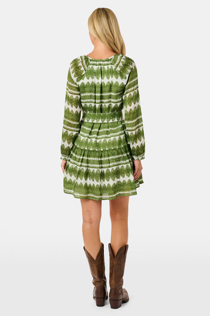A woman outdoors wears the Hayam Monroe Dress by Roller Rabbit—an olive and white long-sleeved dress with a matching belt. She walks with another person, with trees and a wooden fence in the background.