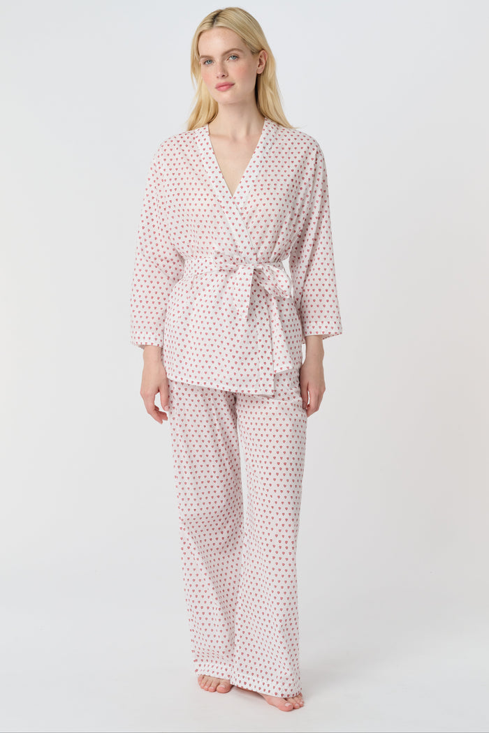 A person stands barefoot in the Roller Rabbit Hearts Christy Set—a light pink lounge set with small red hearts, featuring a belted wrap top and loose-fitting pants—against a plain white background.