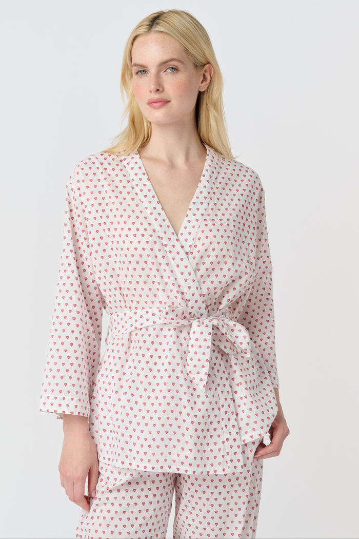 A person stands barefoot in the Roller Rabbit Hearts Christy Set—a light pink lounge set with small red hearts, featuring a belted wrap top and loose-fitting pants—against a plain white background.