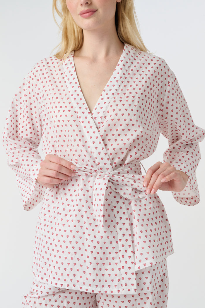 A person stands barefoot in the Roller Rabbit Hearts Christy Set—a light pink lounge set with small red hearts, featuring a belted wrap top and loose-fitting pants—against a plain white background.