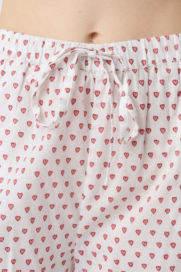 A person stands barefoot in the Roller Rabbit Hearts Christy Set—a light pink lounge set with small red hearts, featuring a belted wrap top and loose-fitting pants—against a plain white background.