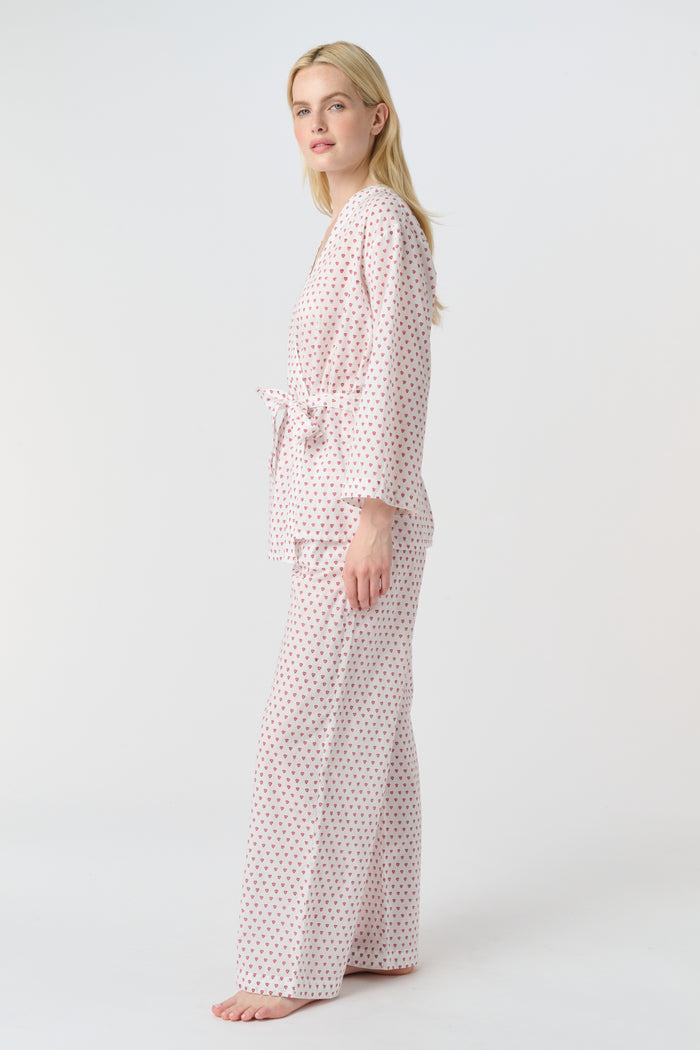 A person stands barefoot in the Roller Rabbit Hearts Christy Set—a light pink lounge set with small red hearts, featuring a belted wrap top and loose-fitting pants—against a plain white background.