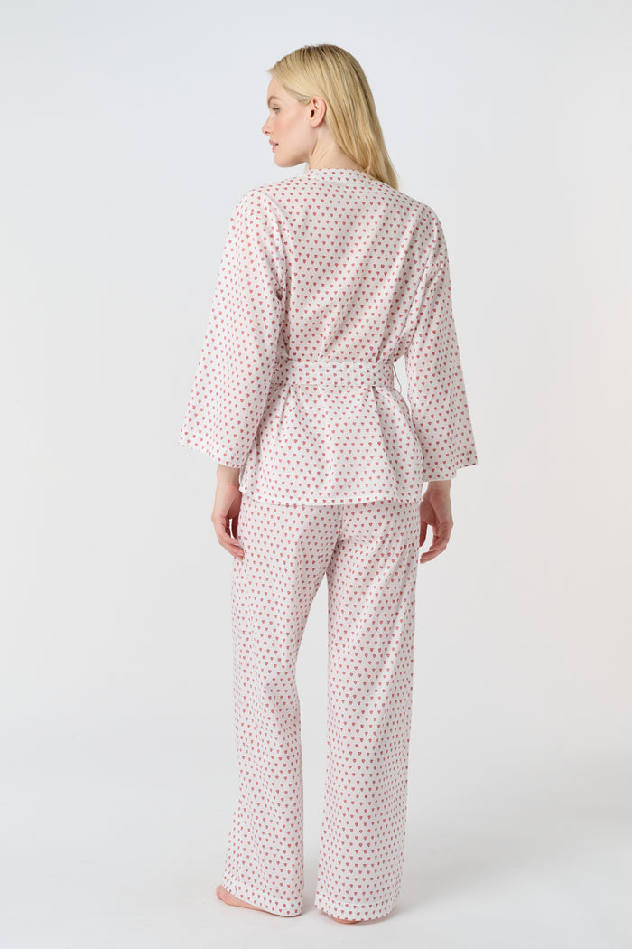 A person stands barefoot in the Roller Rabbit Hearts Christy Set—a light pink lounge set with small red hearts, featuring a belted wrap top and loose-fitting pants—against a plain white background.