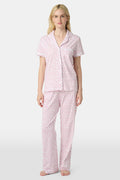 A person stands barefoot in Roller Rabbit’s Hearts Fiora Pajamas—light pink with red hearts. The matching set features a short-sleeve button-up top and long pants, made from soft Pima cotton, against a plain white background.