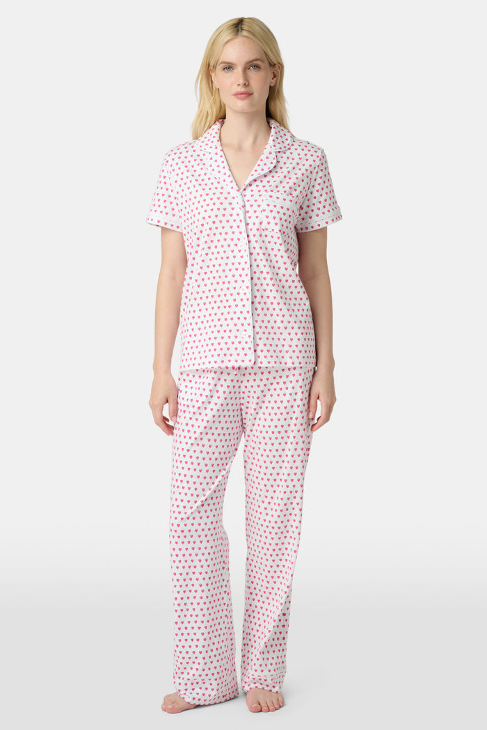 A person stands barefoot in Roller Rabbit’s Hearts Fiora Pajamas—light pink with red hearts. The matching set features a short-sleeve button-up top and long pants, made from soft Pima cotton, against a plain white background.