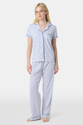 A woman stands barefoot in Roller Rabbit's Hearts Fiora Pajamas, a pima cotton set featuring a white base with small blue and red heart patterns, including a short-sleeve button-up top and elastic waist pants.
