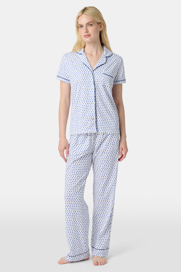 A woman stands barefoot in Roller Rabbit's Hearts Fiora Pajamas, a pima cotton set featuring a white base with small blue and red heart patterns, including a short-sleeve button-up top and elastic waist pants.