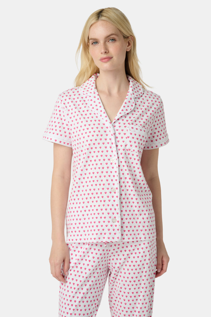 A person stands barefoot in Roller Rabbit’s Hearts Fiora Pajamas—light pink with red hearts. The matching set features a short-sleeve button-up top and long pants, made from soft Pima cotton, against a plain white background.