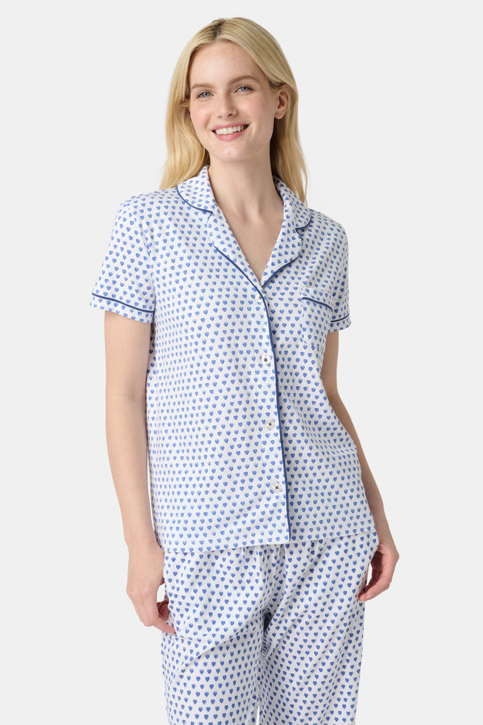 A woman stands barefoot in Roller Rabbit's Hearts Fiora Pajamas, a pima cotton set featuring a white base with small blue and red heart patterns, including a short-sleeve button-up top and elastic waist pants.