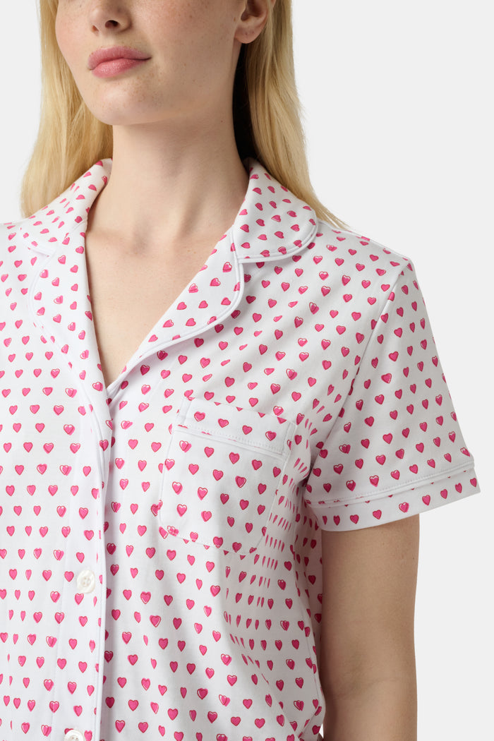 A person stands barefoot in Roller Rabbit’s Hearts Fiora Pajamas—light pink with red hearts. The matching set features a short-sleeve button-up top and long pants, made from soft Pima cotton, against a plain white background.