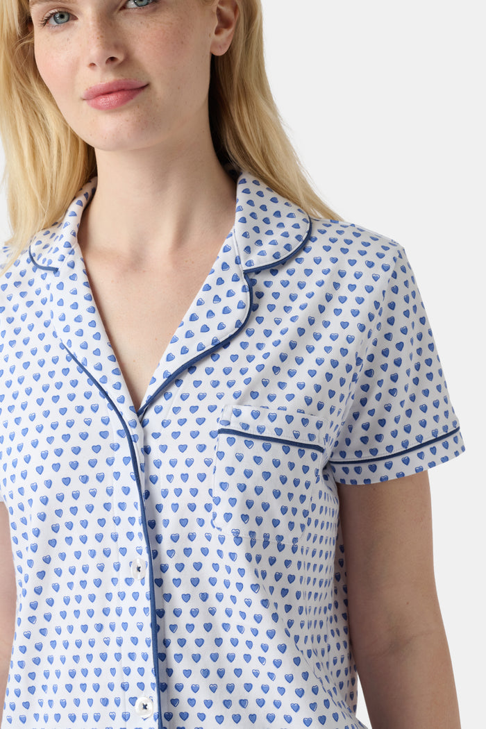 A woman stands barefoot in Roller Rabbit's Hearts Fiora Pajamas, a pima cotton set featuring a white base with small blue and red heart patterns, including a short-sleeve button-up top and elastic waist pants.