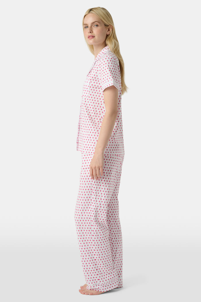 A person stands barefoot in Roller Rabbit’s Hearts Fiora Pajamas—light pink with red hearts. The matching set features a short-sleeve button-up top and long pants, made from soft Pima cotton, against a plain white background.
