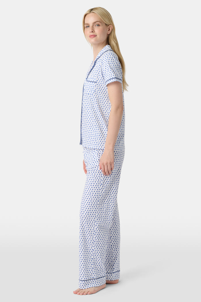 A woman stands barefoot in Roller Rabbit's Hearts Fiora Pajamas, a pima cotton set featuring a white base with small blue and red heart patterns, including a short-sleeve button-up top and elastic waist pants.