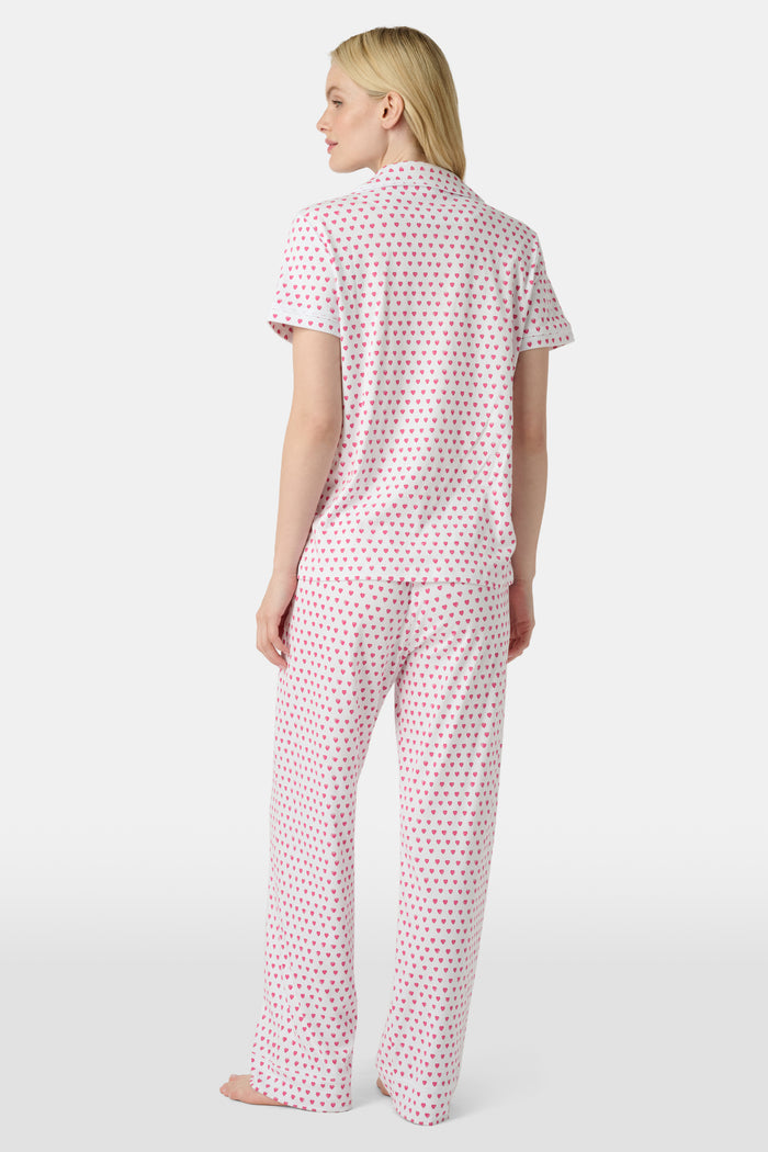 A person stands barefoot in Roller Rabbit’s Hearts Fiora Pajamas—light pink with red hearts. The matching set features a short-sleeve button-up top and long pants, made from soft Pima cotton, against a plain white background.
