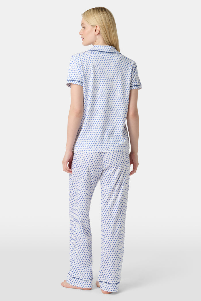 A woman stands barefoot in Roller Rabbit's Hearts Fiora Pajamas, a pima cotton set featuring a white base with small blue and red heart patterns, including a short-sleeve button-up top and elastic waist pants.