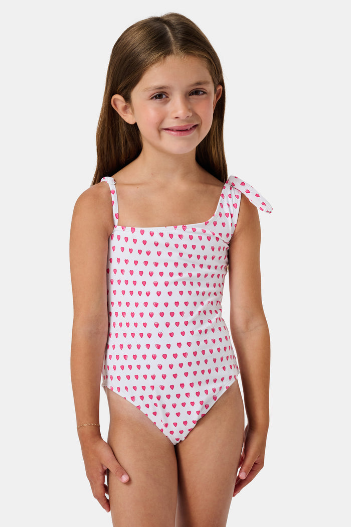 The Roller Rabbit Hearts Kids Jacey One Piece is a swimsuit with an asymmetrical neckline, white fabric dotted with pink hearts, and an adjustable knot-tied shoulder strap.