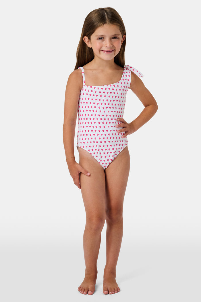 The Roller Rabbit Hearts Kids Jacey One Piece is a swimsuit with an asymmetrical neckline, white fabric dotted with pink hearts, and an adjustable knot-tied shoulder strap.
