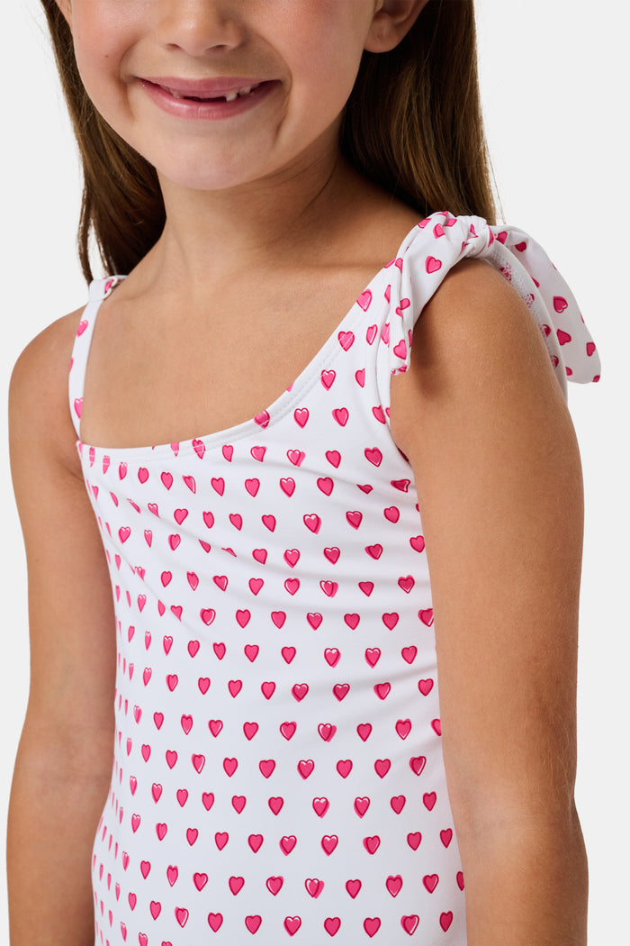 The Roller Rabbit Hearts Kids Jacey One Piece is a swimsuit with an asymmetrical neckline, white fabric dotted with pink hearts, and an adjustable knot-tied shoulder strap.
