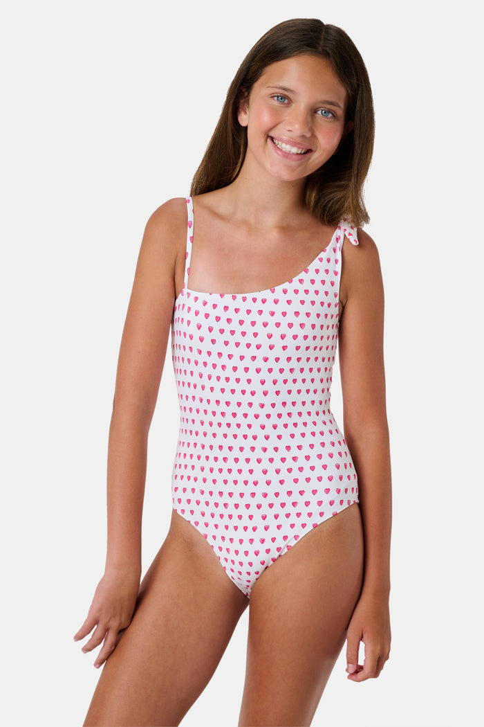 The Roller Rabbit Hearts Kids Jacey One Piece is a swimsuit with an asymmetrical neckline, white fabric dotted with pink hearts, and an adjustable knot-tied shoulder strap.