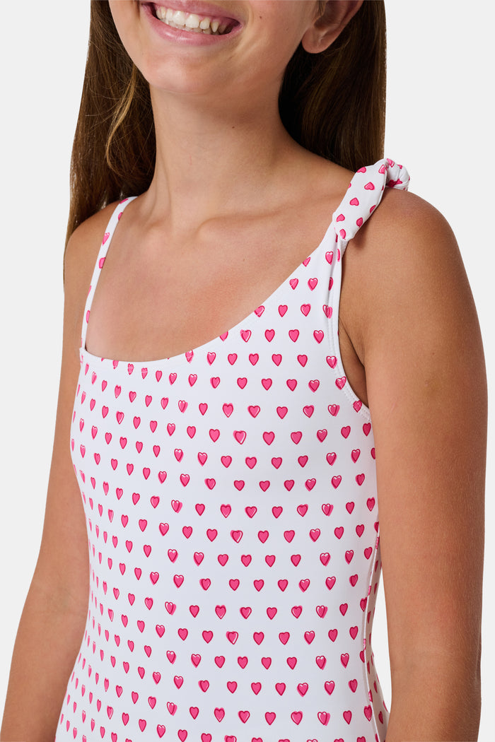 The Roller Rabbit Hearts Kids Jacey One Piece is a swimsuit with an asymmetrical neckline, white fabric dotted with pink hearts, and an adjustable knot-tied shoulder strap.
