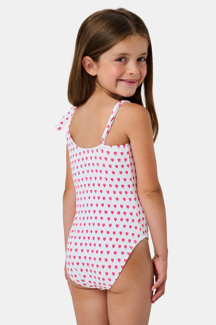 The Roller Rabbit Hearts Kids Jacey One Piece is a swimsuit with an asymmetrical neckline, white fabric dotted with pink hearts, and an adjustable knot-tied shoulder strap.