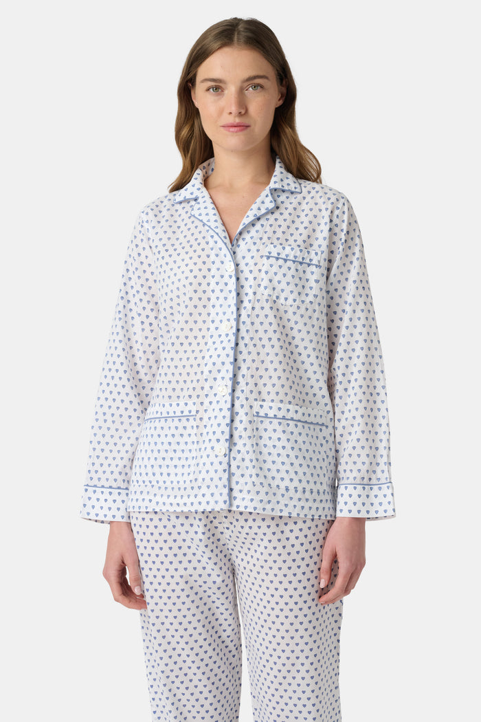 A person stands barefoot in Roller Rabbit’s Hearts Loungewear, a matching cotton pajama set with heart prints, featuring a button-up long sleeve top and long pants. The background is plain white.